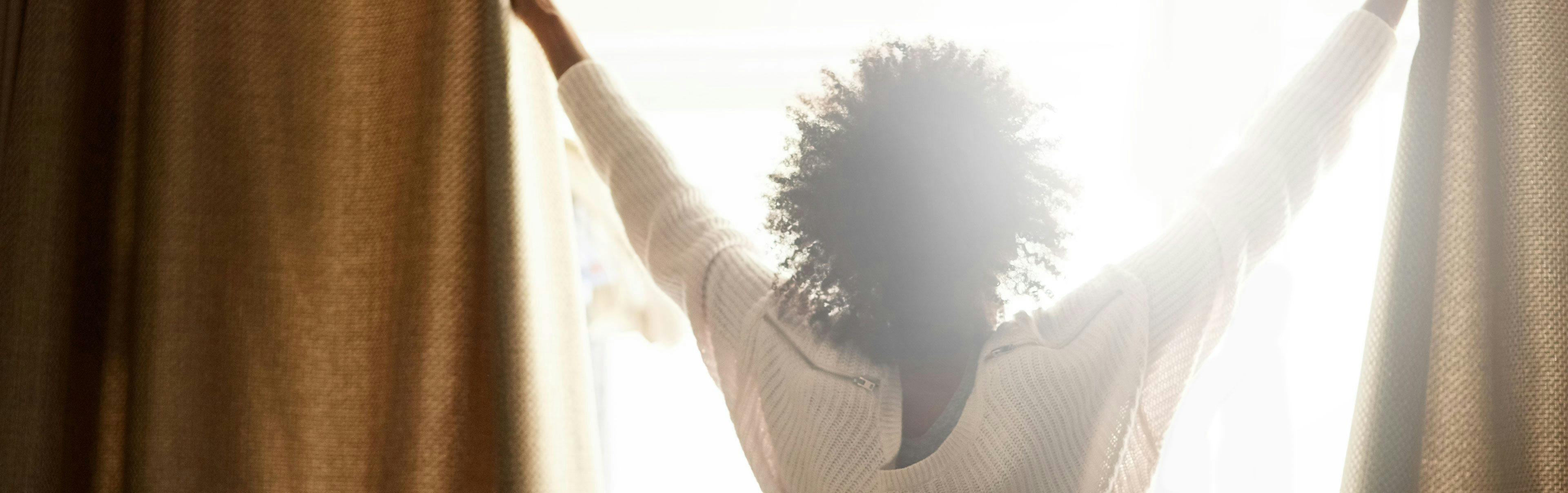 A women opening her curtain to let the sun shine into her room.