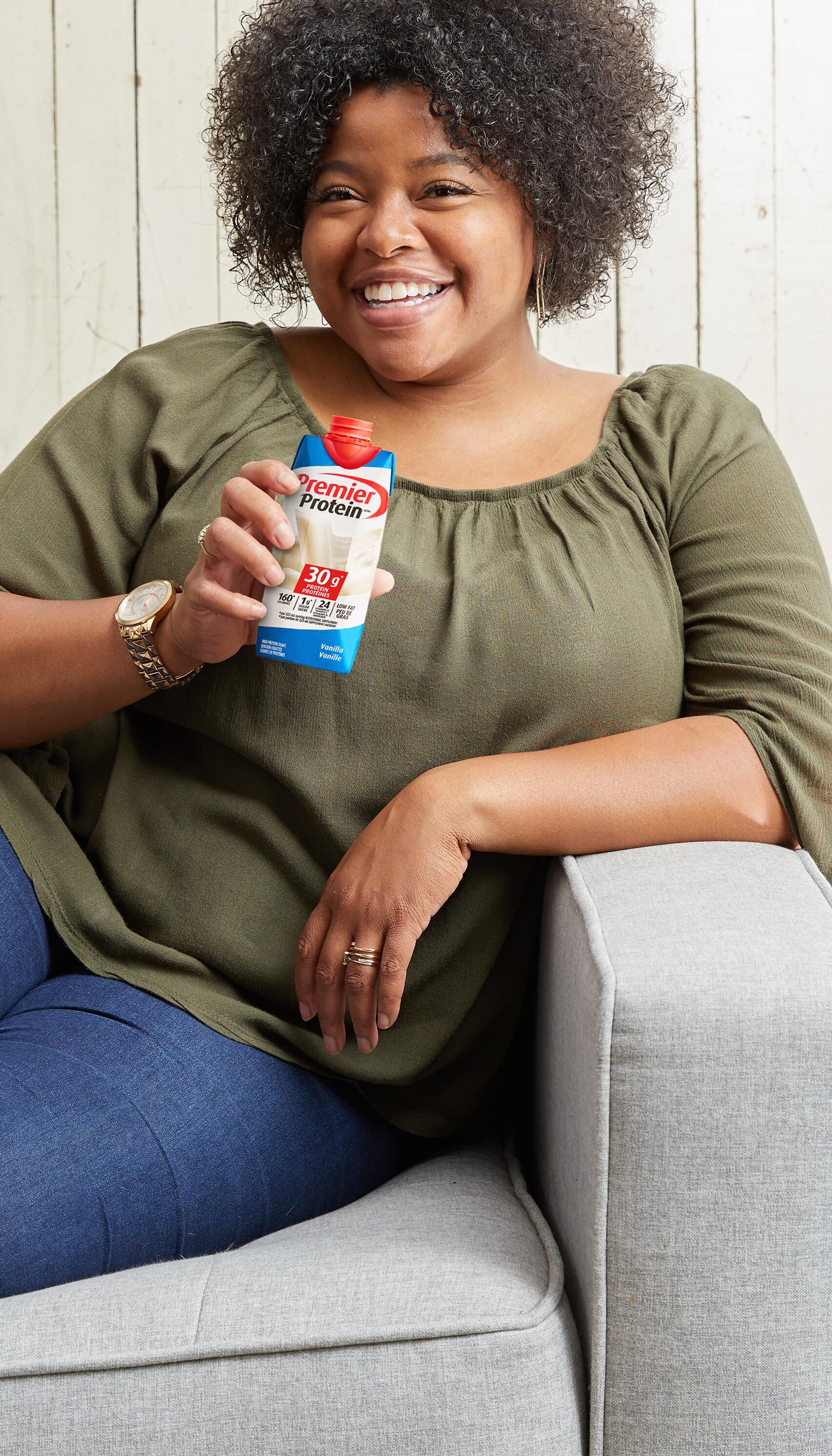 A smiling woman relaxing on a sofa while holding a Premier Protein Vanilla shake.