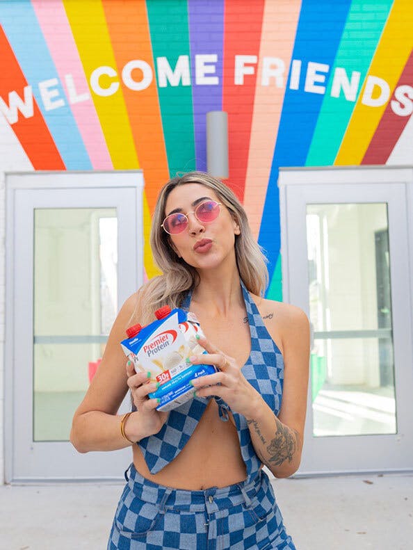 Woman holding a 4 pack of Premier Protein shakes in front of a colorful mural that reads 'welcome friends'