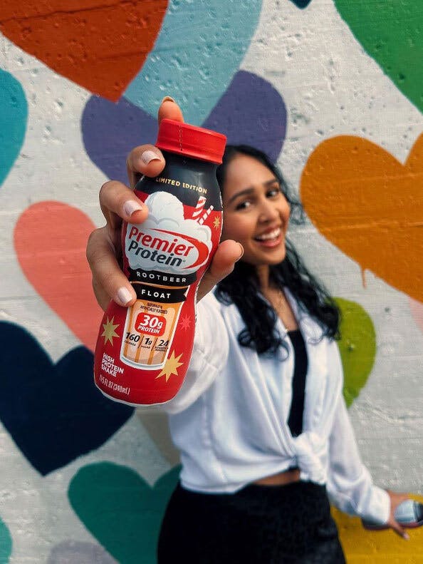 Woman holding up a salted caramel popcorn shake standing in front of a mural of colorful hearts
