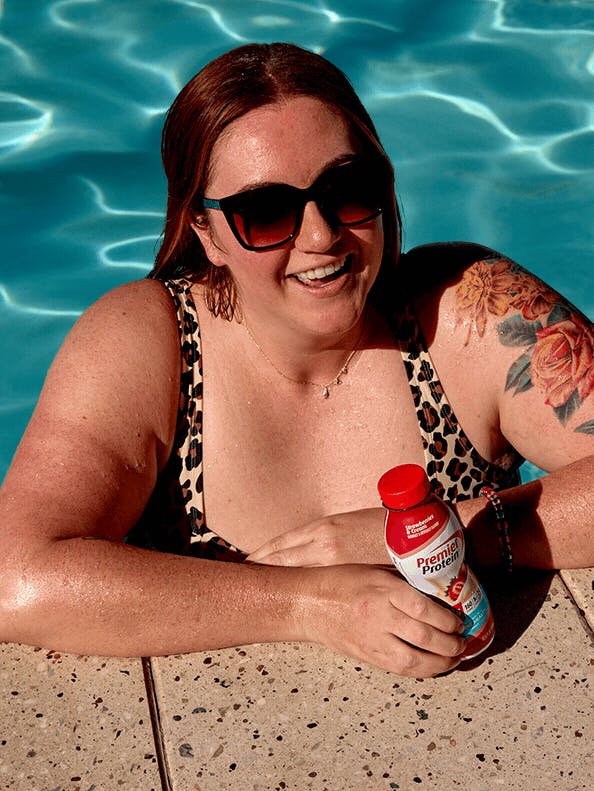A swimmer in sunglasses smiles while holding a premier protein shake at the edge of the pool. The pool water is animated.