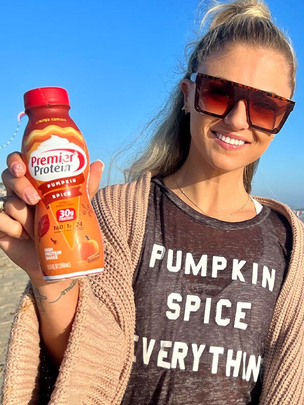 A woman wearing a shirt reading 'pumpkin spice everything' while holding up a pumpkin spice flavored protein shake