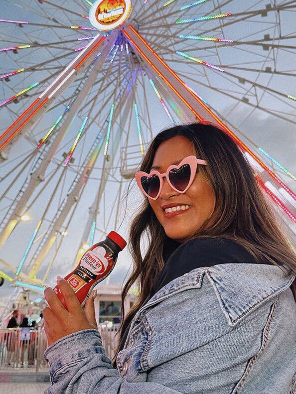Woman wearing heart shaped glasses smiles with a premier protein shake in front of a ferris wheel