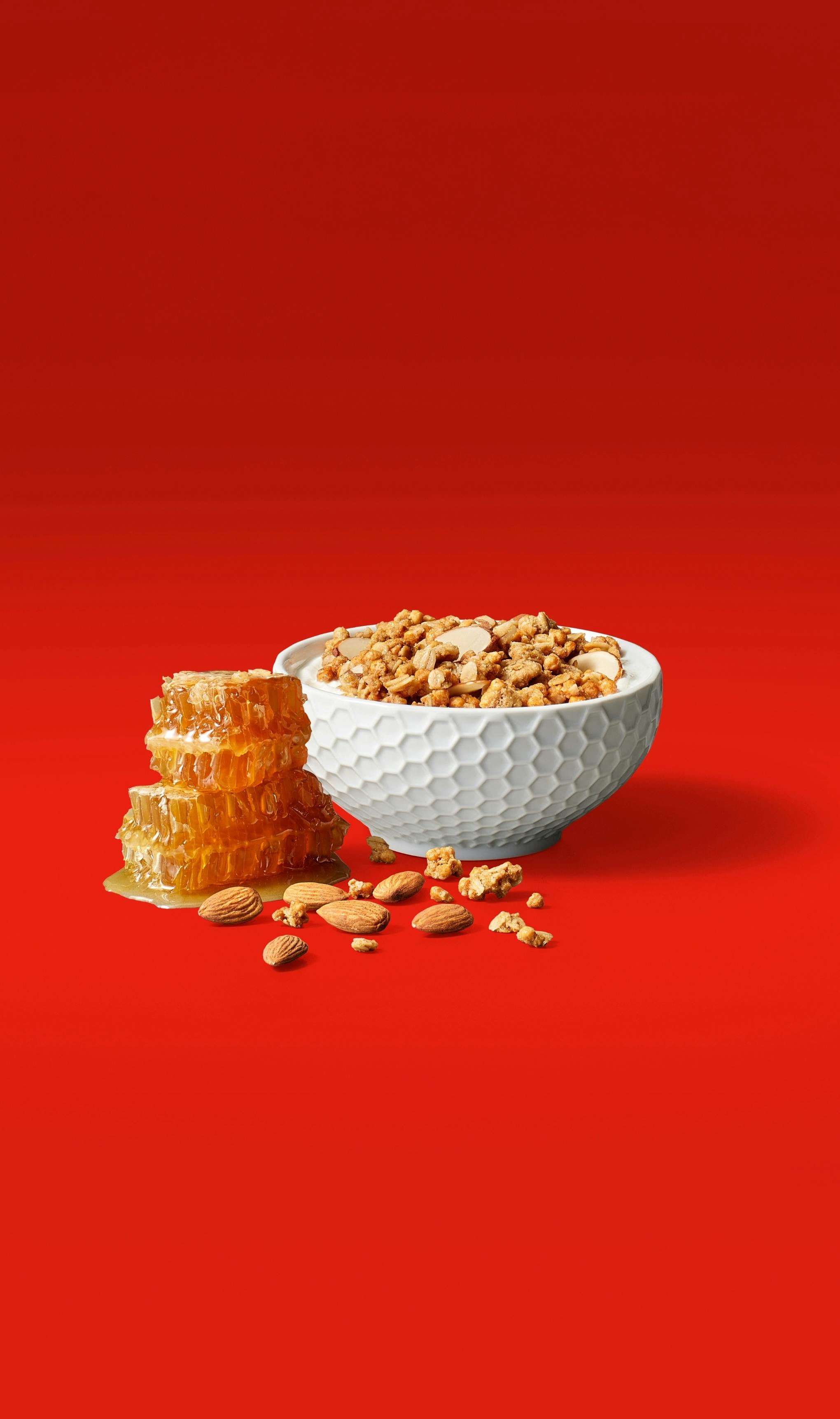 A bag of Premier Protein Honey Almond Protein Granola behind a bowl of granola and a stack of honeycombs and almonds.